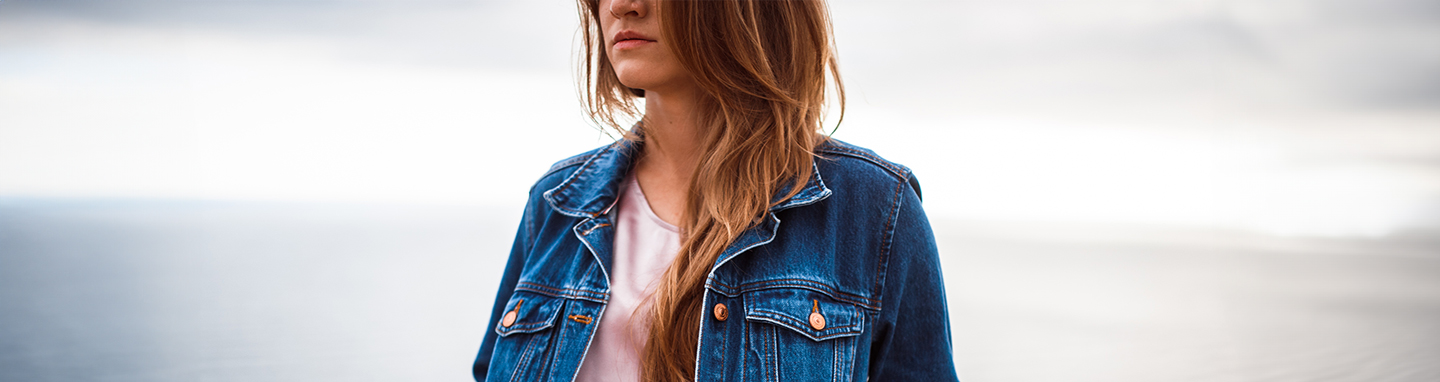 Woman in a denim jacket and jeans poses on a cliff in front of the ocean.