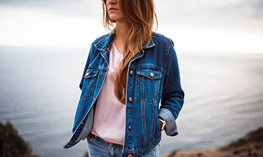 Woman in a denim jacket and jeans poses on a cliff in front of the ocean.